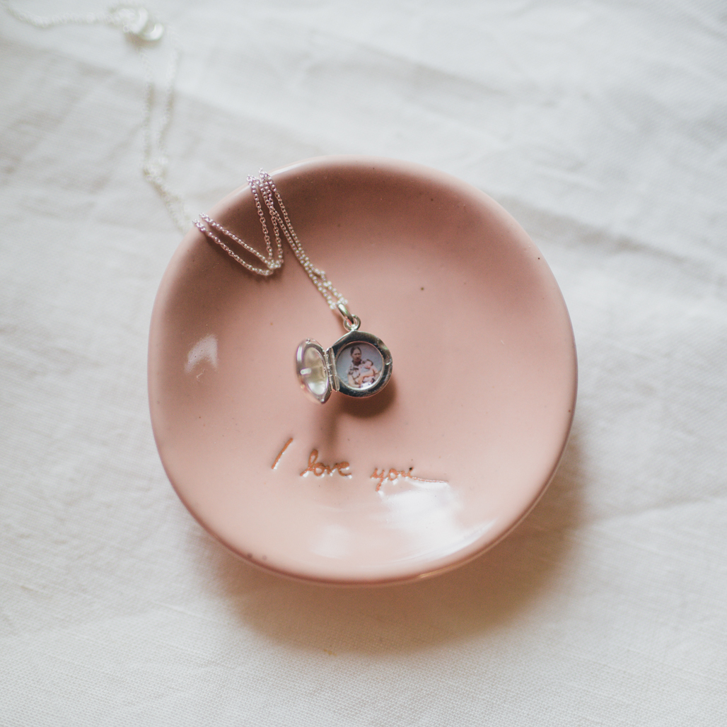 very small silver photo locket necklace on pink jewelry dish with image of her mother holding her when she was a baby